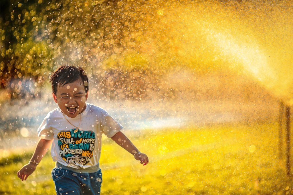A young boy in a T-shirt and jeans running through sprinklers outside.